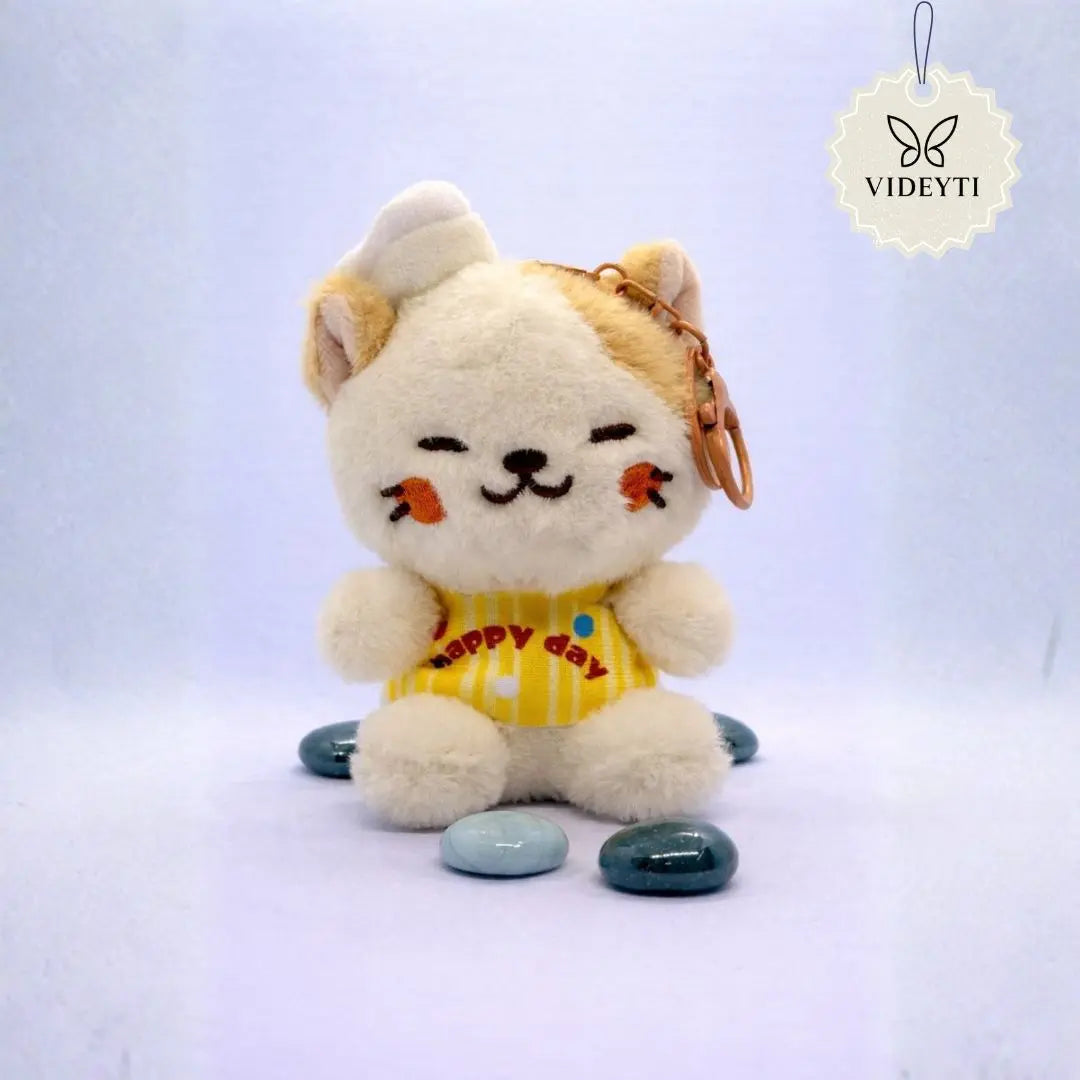 Cream plush cat bag charm wearing a yellow Happy Day outfit with embroidered details and a rose-gold keychain ring.