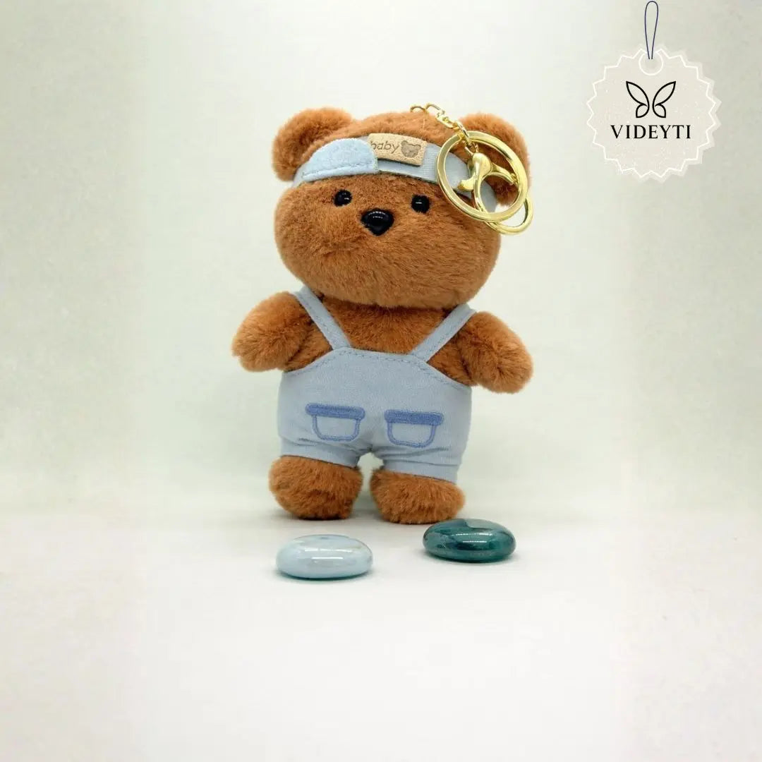 Cream plush teddy bear bag charm wearing brown overalls and a matching brown cap with a gold keychain ring.
