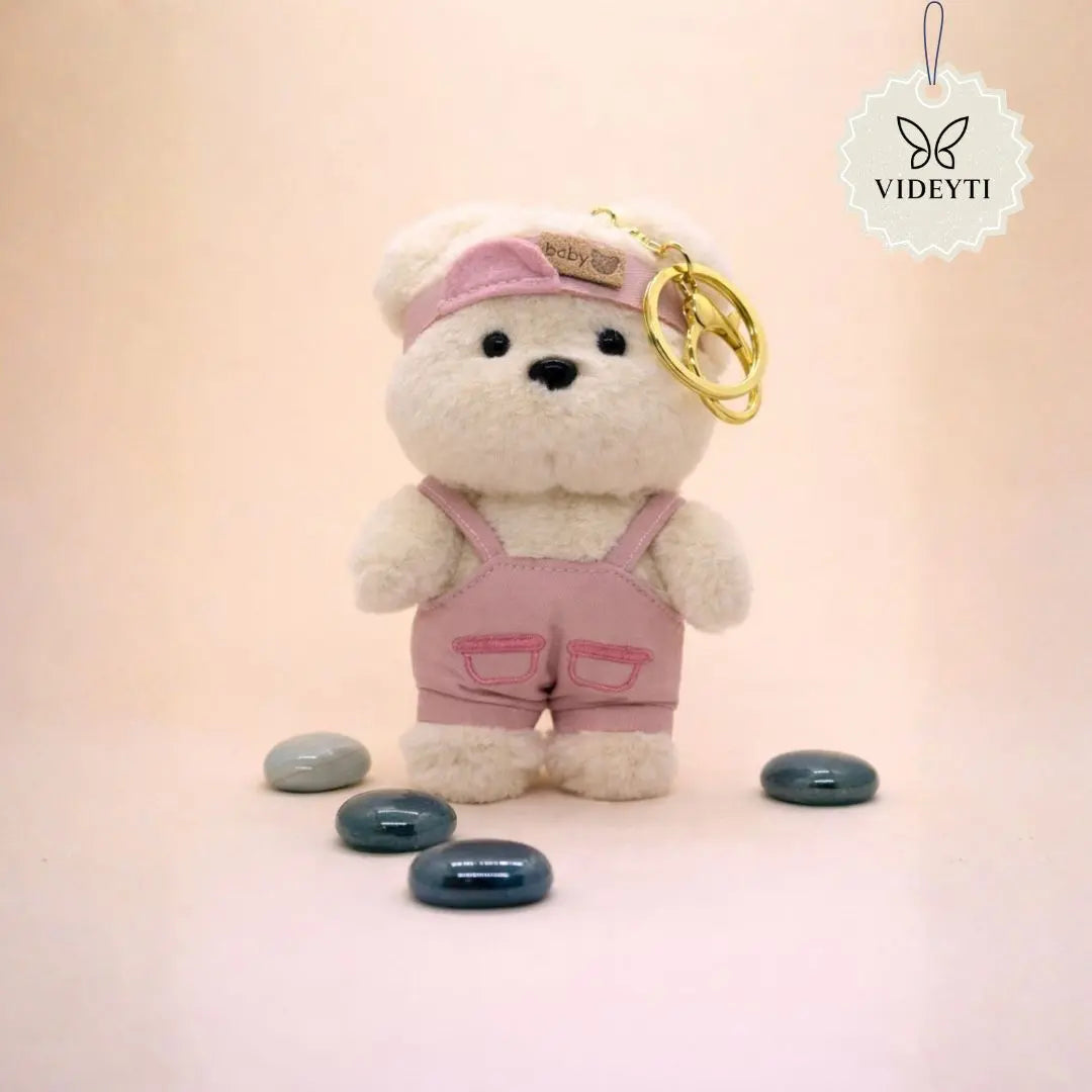 Cream plush teddy bear bag charm wearing pink overalls and matching cap with a gold keychain ring.