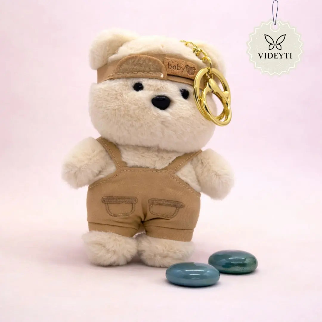 Brown plush teddy bear bag charm wearing blue overalls and a matching blue cap with a gold keychain ring.
